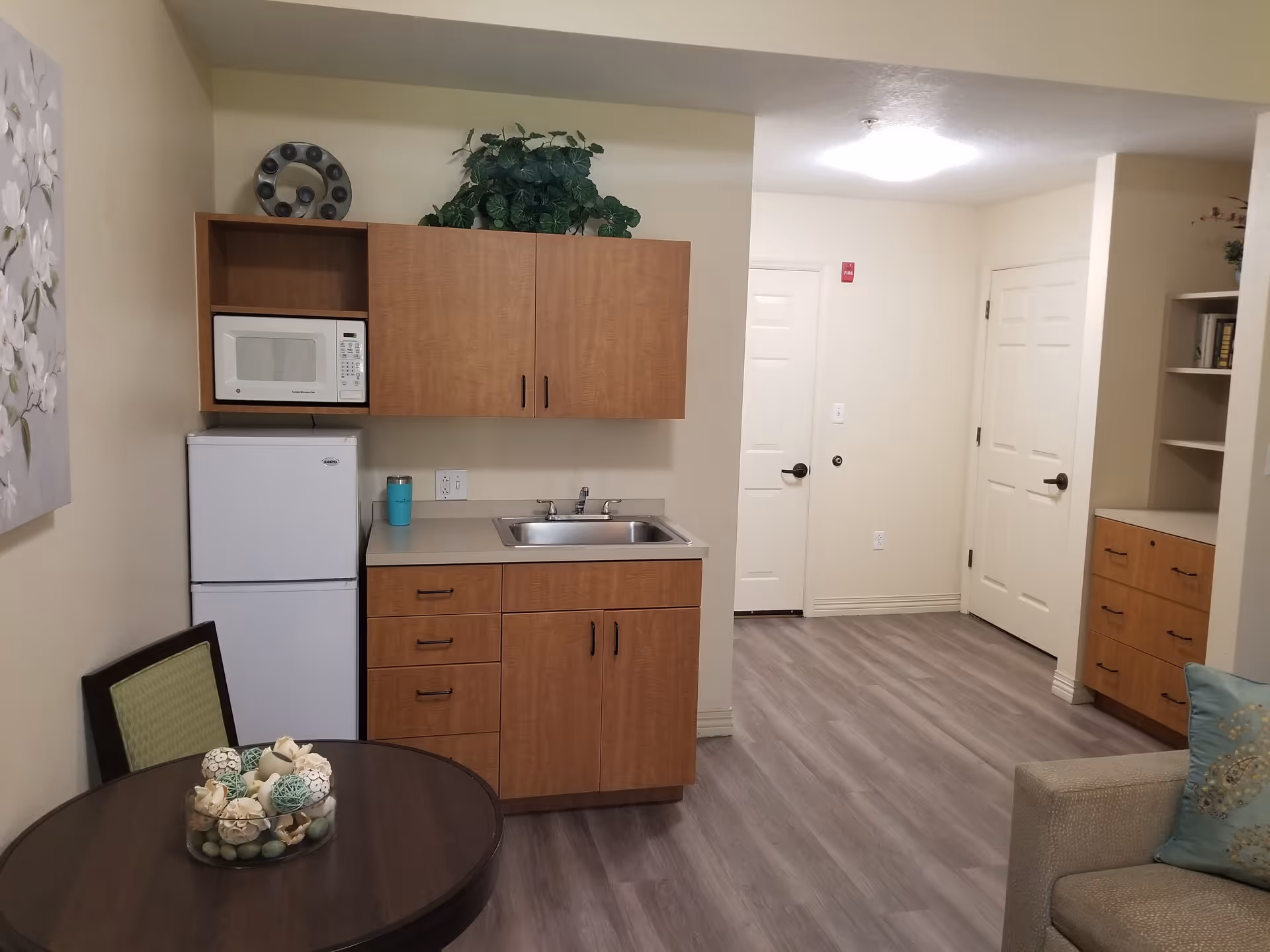 Interior view of a small kitchen area in a senior living facility with wooden cabinets, a white microwave, a white refrigerator, a sink, and a round table with a decorative centerpiece. The room has light-colored walls and flooring, with a hallway and two closed white doors visible in the background.