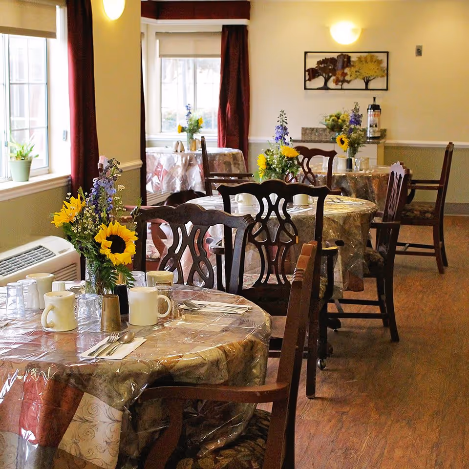 Dining room with round tables covered in plastic tablecloths, each set with white mugs, utensils, and floral centerpieces featuring sunflowers and other flowers. Wooden chairs surround the tables, and windows with red curtains allow natural light into the room.