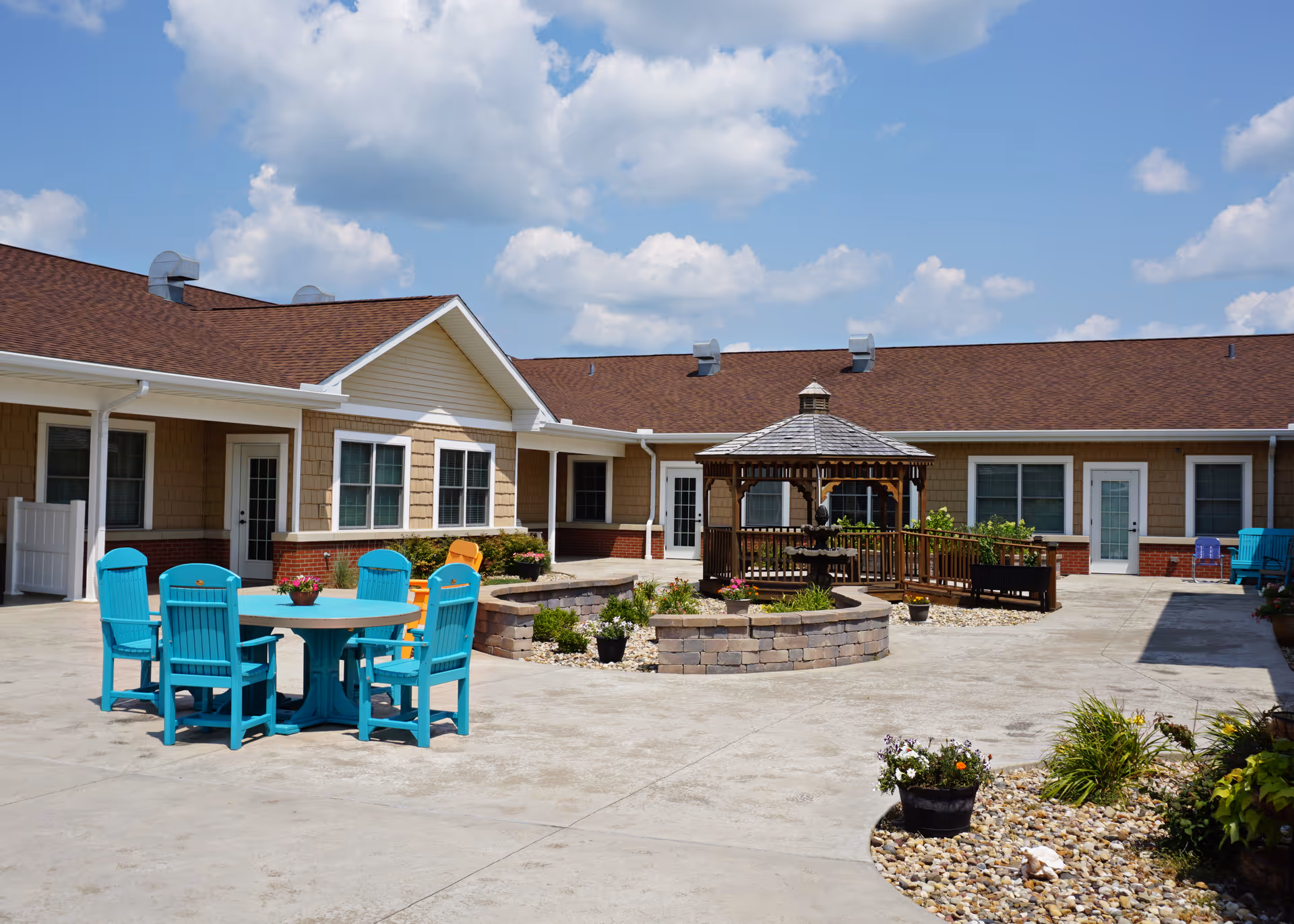 Outdoor courtyard area of Snyder Village Assisted Living featuring a round table with turquoise chairs, a wooden gazebo with a fountain inside, potted plants, and a building with beige siding and brown roof under a partly cloudy sky.