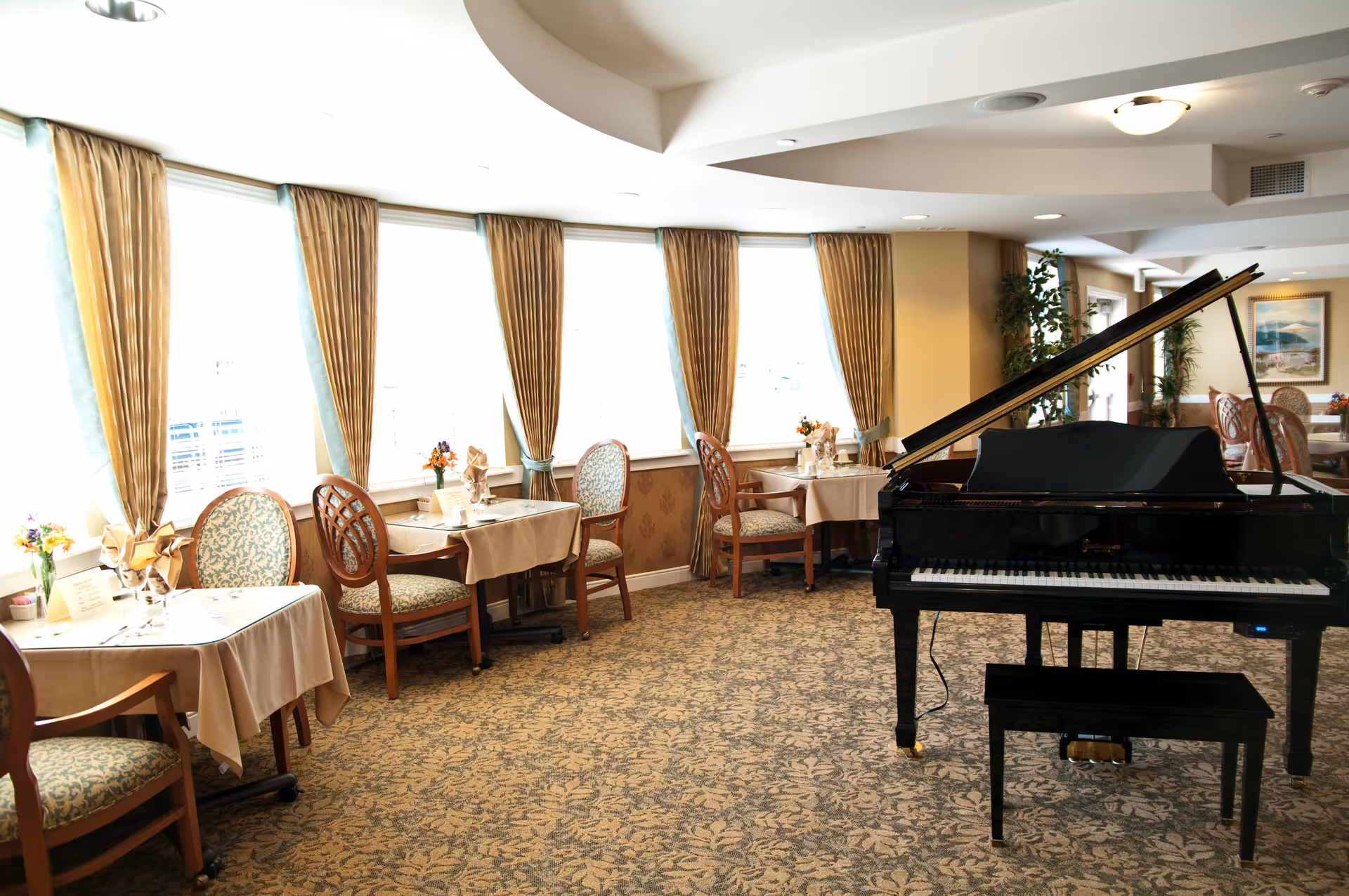 A bright dining room with several tables covered in beige tablecloths, each set with chairs and small floral centerpieces. Large windows with gold curtains allow natural light to fill the room. A black grand piano with a matching bench is positioned prominently in the foreground.