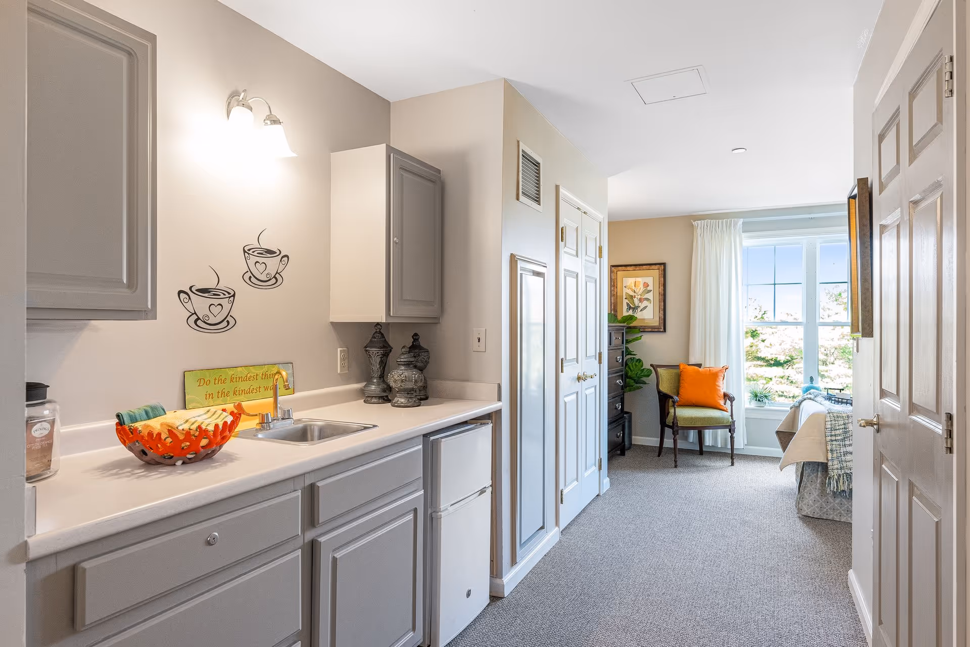 A bright and cozy interior space featuring a small kitchenette with gray cabinets, a sink, and a mini fridge on the left. The wall above the counter has two coffee cup decals and a sign that reads 'Do the kindest thing in the kindest way.' The room extends into a sitting area with a green chair adorned with an orange pillow, a framed floral picture on the wall, a chest of drawers, and a large window letting in natural light. The floor is carpeted and the walls are painted in neutral tones.