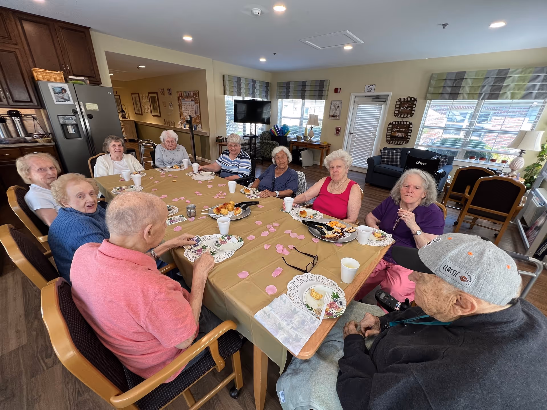 A group of elderly residents seated around a decorated table eating and socializing in a senior living facility common dining area.