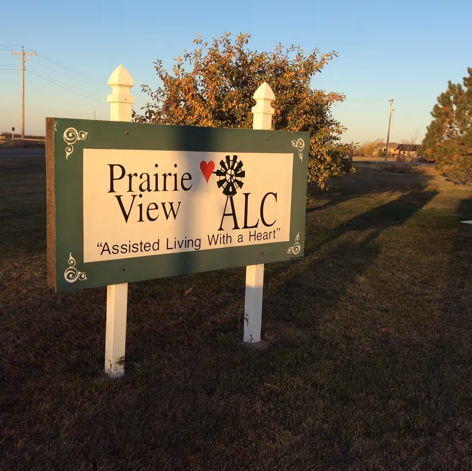 A green and white outdoor sign for Prairie View ALC with the slogan 'Assisted Living With a Heart' in a grassy area with trees and a clear sky at sunset.