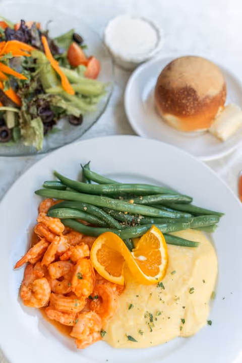 A plate of cooked shrimp, green beans, and creamy polenta garnished with two orange slices. Next to the plate is a small bread roll with a pat of butter on a white saucer, a glass of milk, and a side salad with mixed greens, cherry tomatoes, sliced black olives, and shredded carrots.
