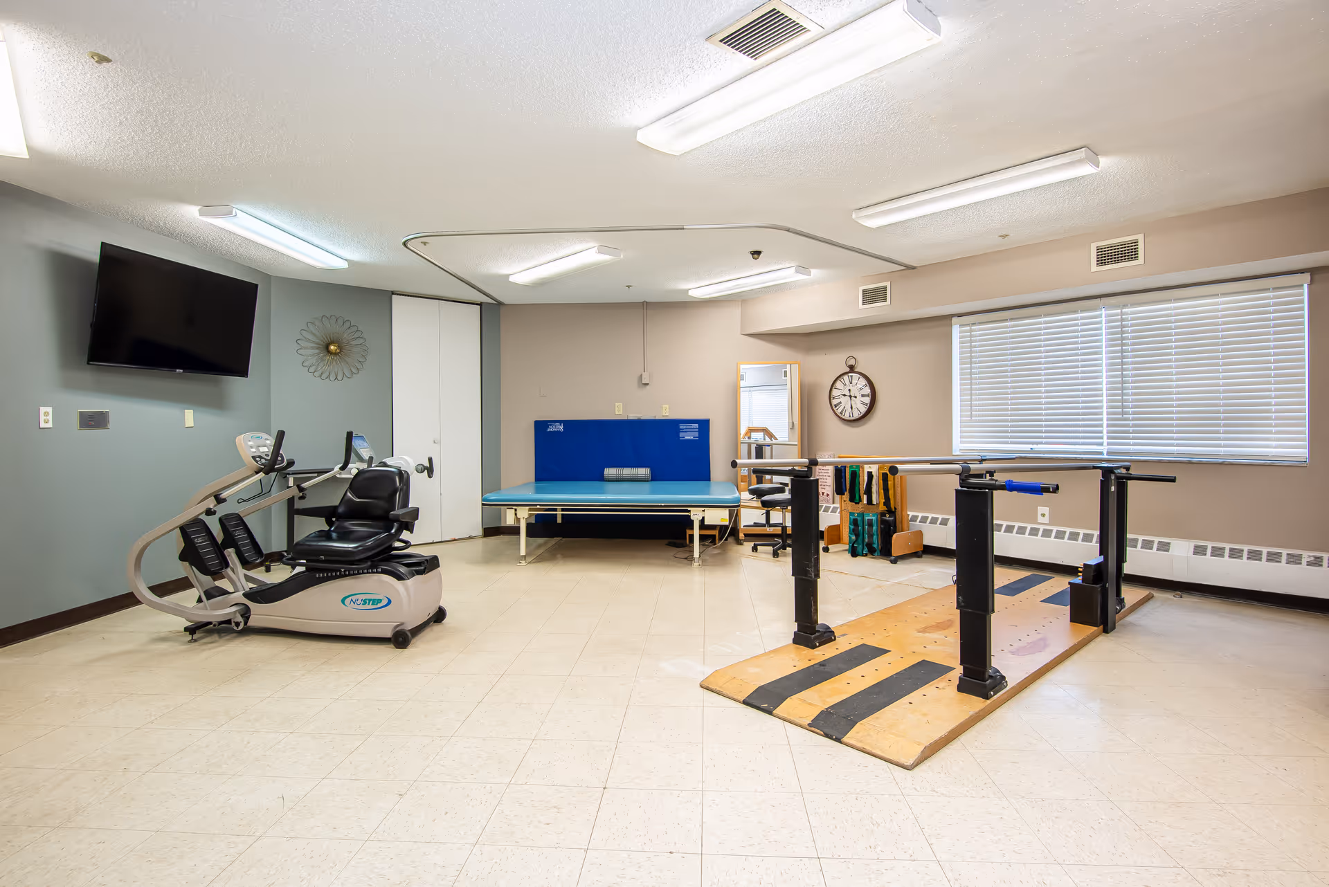 A rehabilitation or physical therapy room with exercise equipment including a recumbent bike, parallel bars for walking practice, a padded therapy table, a wall clock, and a large window with blinds.