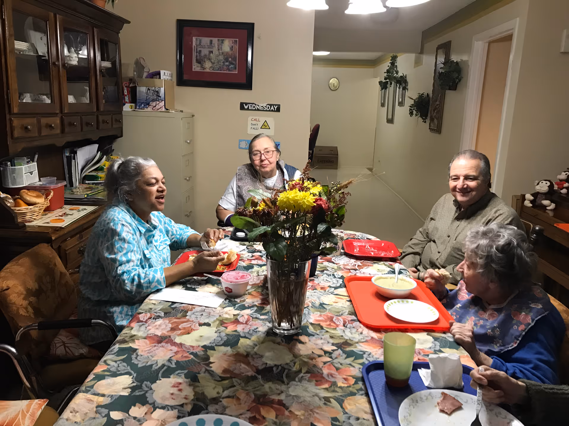 Four elderly people sitting around a dining table with a floral tablecloth, eating a meal together in a cozy room. There is a vase with yellow and red flowers in the center of the table. The room has wooden furniture, including a cabinet with dishes and a filing cabinet in the background. The walls are decorated with framed pictures and plants.