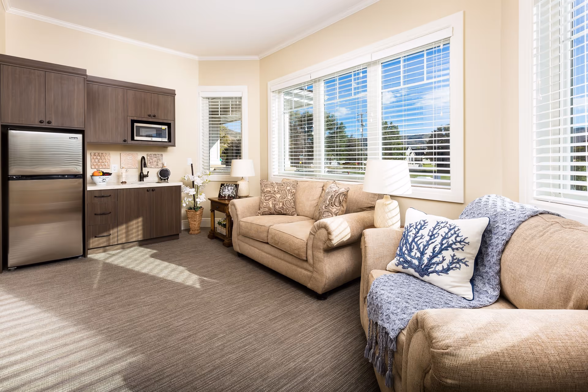 A bright and cozy living area in a senior living facility featuring two beige sofas with decorative pillows and a blue throw blanket. The room has large windows with white blinds letting in natural light. There is a small wooden side table with a lamp and framed photo, and a kitchenette with dark wood cabinets, a stainless steel refrigerator, microwave, and a sink. The walls are painted light beige and the floor is carpeted.