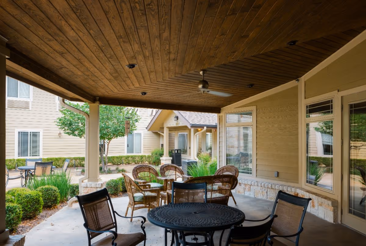 Covered outdoor patio area with a wooden ceiling and ceiling fan, featuring a round metal table with four chairs in the foreground and a set of wicker chairs with cushions around a small table in the background. The patio overlooks a landscaped courtyard with bushes, trees, and beige building walls with windows.