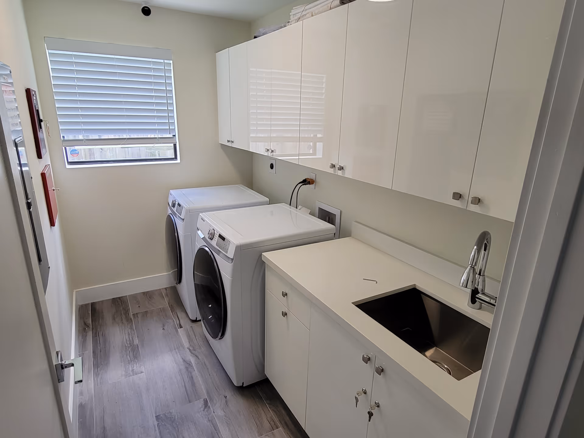A clean and modern laundry room with a front-loading washing machine and dryer side by side. Above the machines are white cabinets for storage. To the right is a countertop with a stainless steel sink and a modern faucet. A window with closed blinds is on the left wall, allowing natural light into the room. The floor has wood-like tiles.