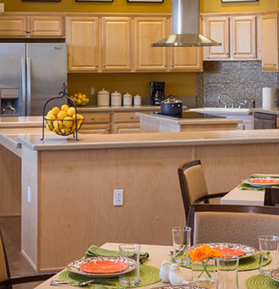 A modern kitchen and dining area with light wood cabinets, a stainless steel refrigerator, and a kitchen island with a stovetop and a pot. The dining table is set with plates, glasses, green placemats, and a small orange flower centerpiece. A basket of lemons is on the kitchen counter.