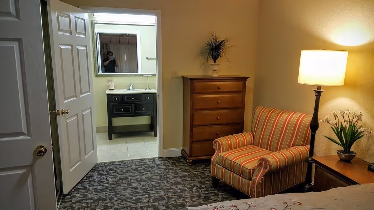 Interior view of a senior living facility room showing a striped armchair next to a wooden chest of drawers and a floor lamp. The room has carpeted flooring and beige walls. An open door reveals a bathroom with a black vanity, a sink, a mirror, and tiled flooring. A person is visible in the bathroom mirror taking the photo.