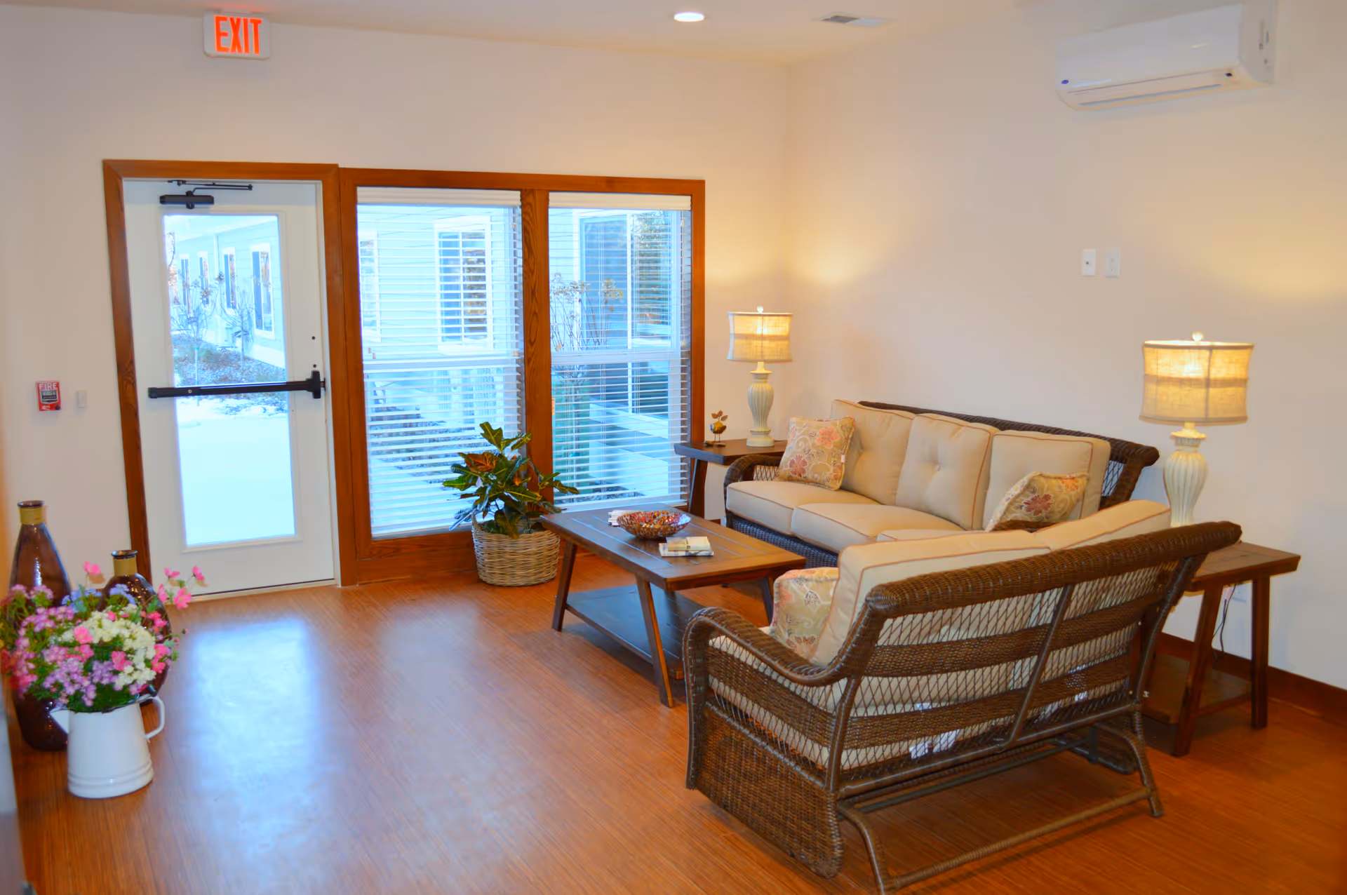 A cozy seating area in a senior living facility with a wicker sofa and loveseat featuring beige cushions and floral pillows. There are two wooden side tables with matching lamps on either side of the sofa, a wooden coffee table in the center, and a potted plant near a glass door and window. The floor is wooden, and the walls are light-colored with an air conditioning unit mounted above.