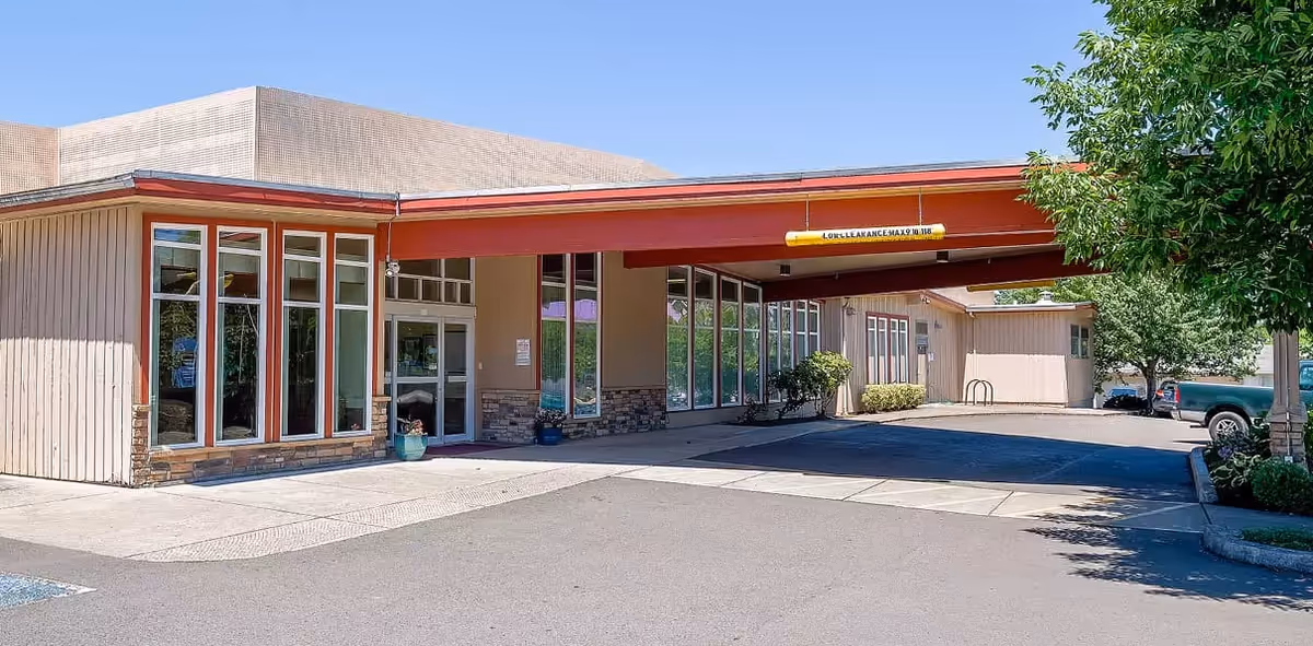 Exterior view of Avamere Rehabilitation of Eugene showing the entrance with large windows, a covered drop-off area with a red roof, and surrounding trees and parked vehicles under a clear blue sky.