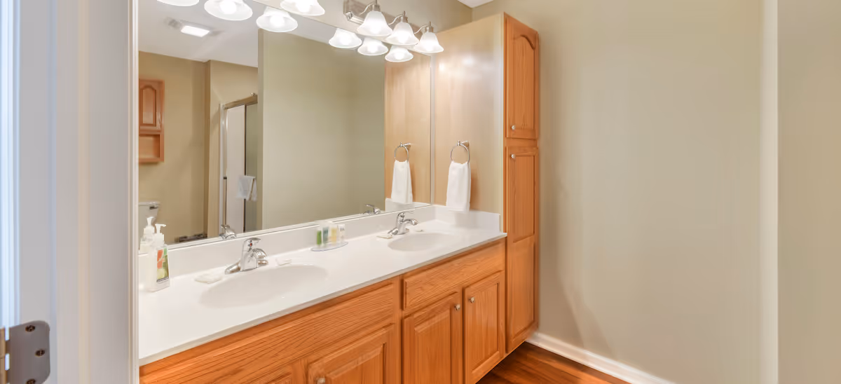 A bathroom with a double sink vanity featuring wooden cabinets and a large mirror above. There are two towel rings with white towels, soap dispensers, and toiletries on the countertop. In the background, a shower with a glass door and a toilet are visible. The floor is wooden and the walls are painted beige.