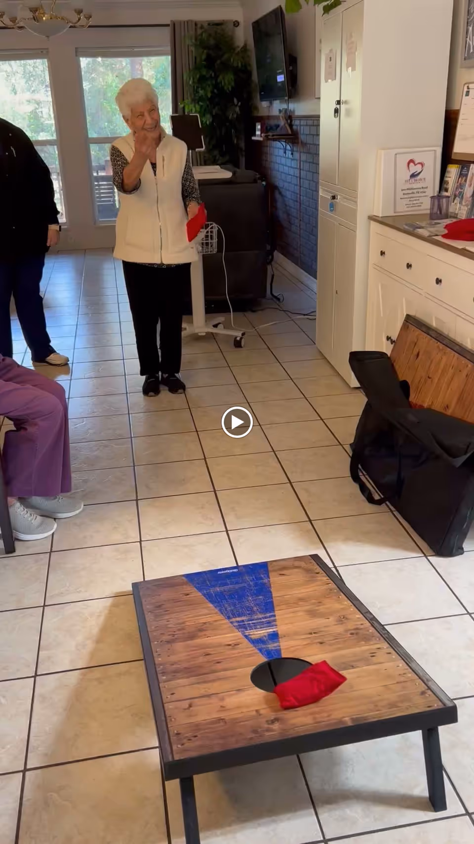 An elderly woman standing indoors on a tiled floor, smiling and holding a red bean bag, playing a bean bag toss game with a wooden board that has a hole and a blue triangle design. Other people are partially visible in the room, which has large windows, a TV mounted on the wall, and cabinets.