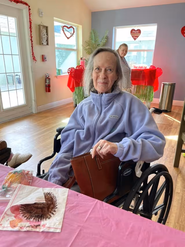 An elderly woman in a wheelchair wearing a light purple jacket sits at a table covered with a pink tablecloth. Behind her, another woman stands near a window decorated with Valentine's Day heart decorations. The room has wooden floors and light-colored walls with festive red and pink decorations.