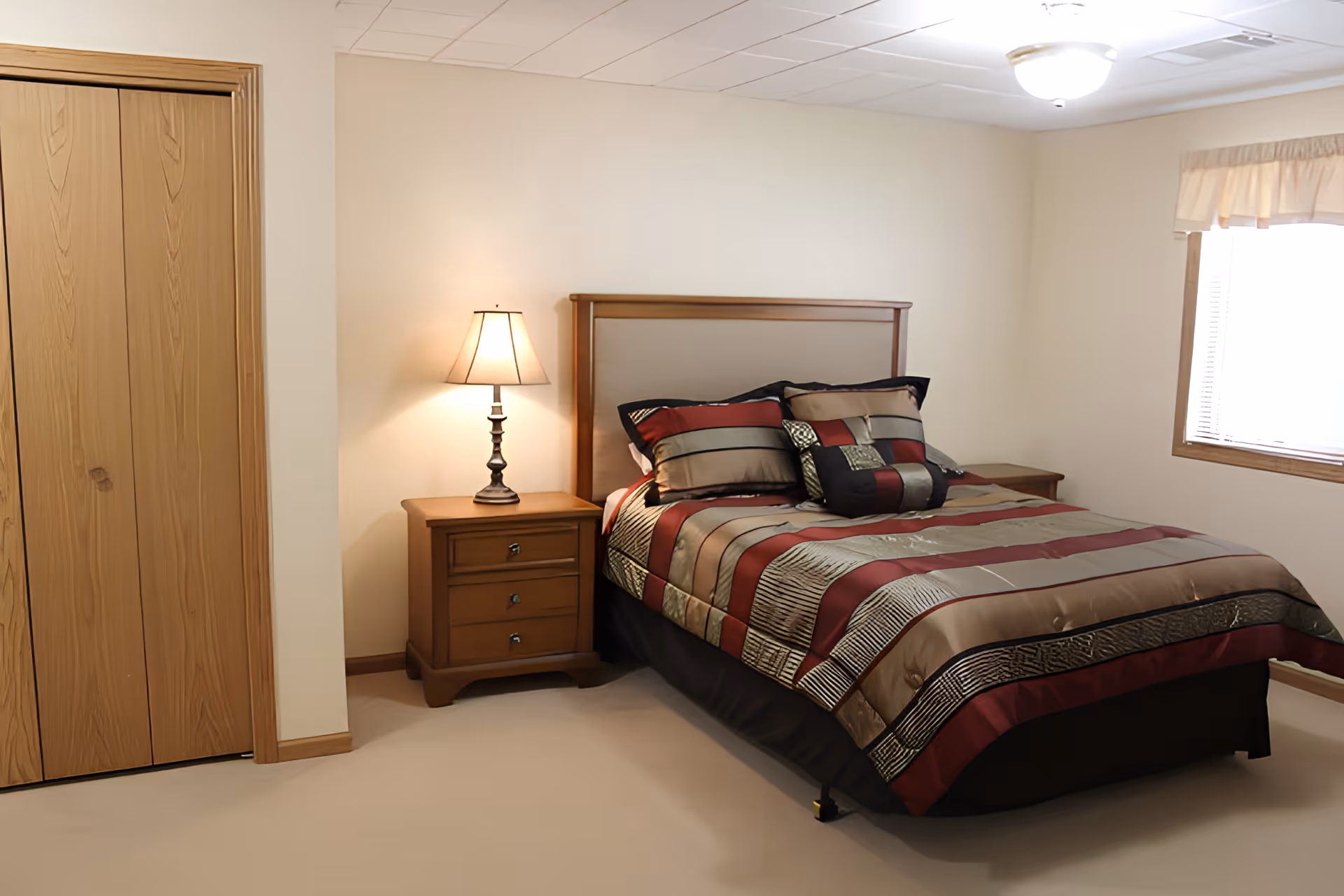 A simple furnished bedroom with a patterned bed, wooden headboard, nightstand with a lamp, a closet, and a window.