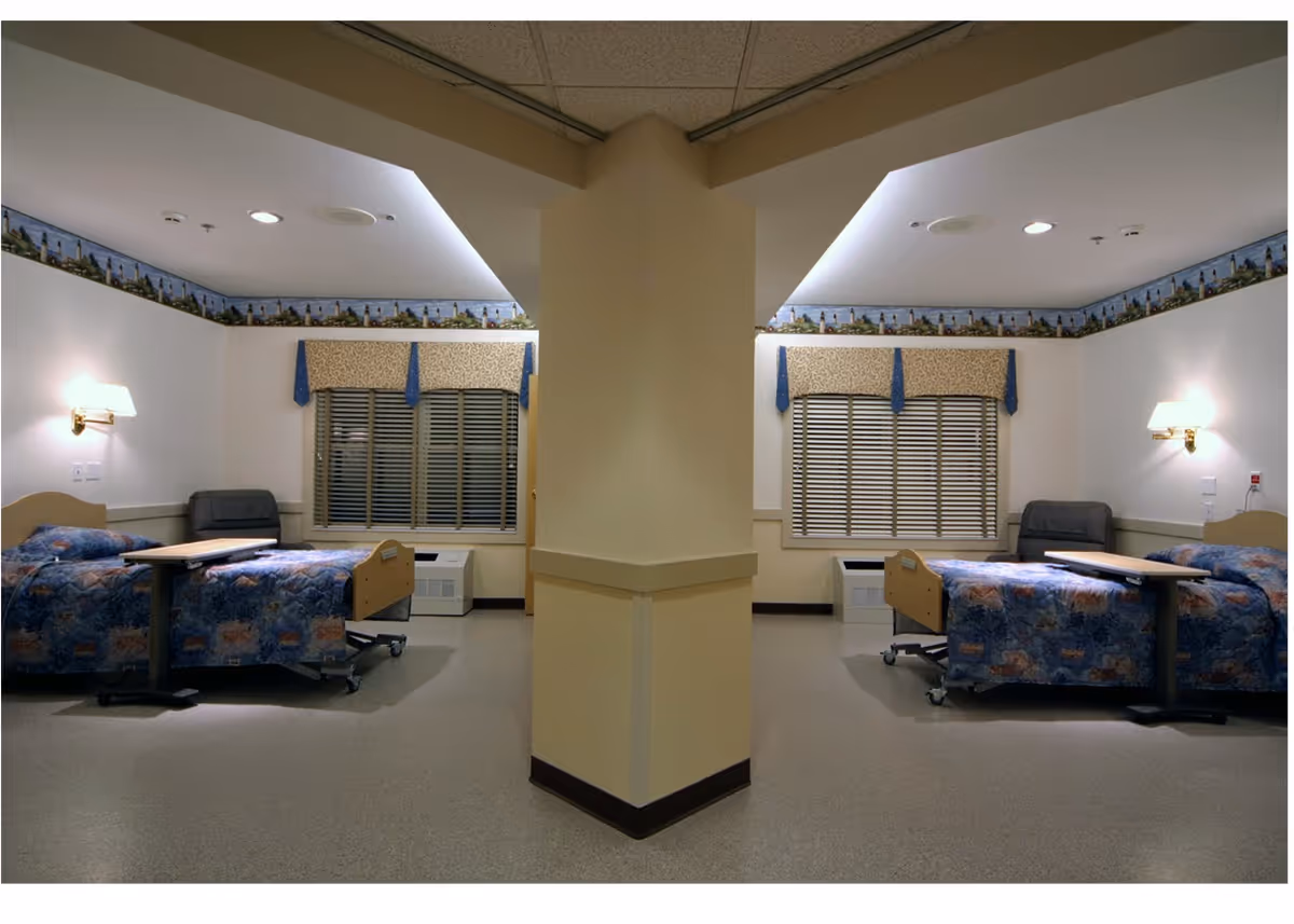 Symmetrical senior-care room with two hospital-style beds, overbed tables, armchairs and a central pillar.