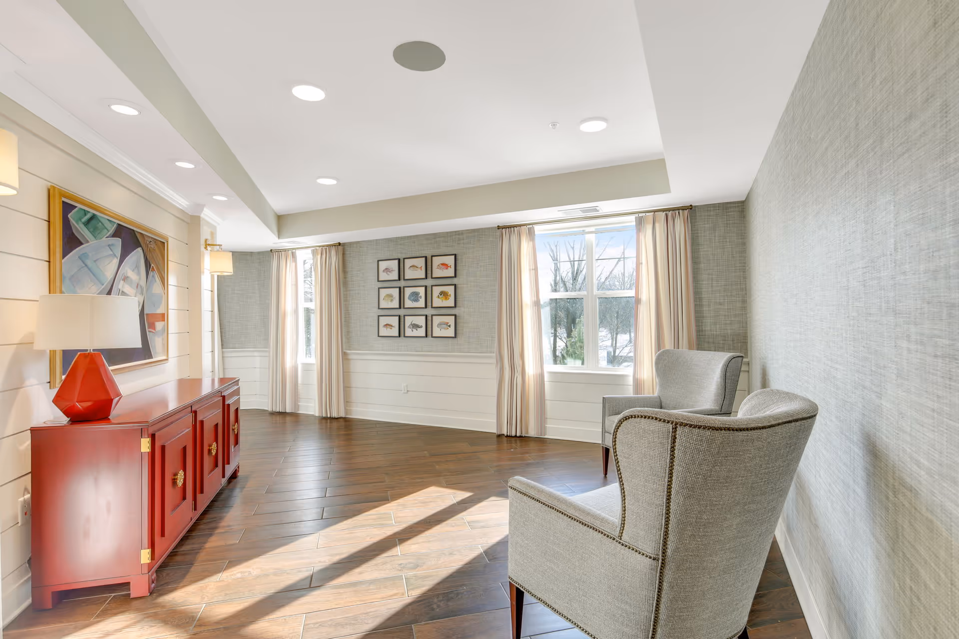 Sunlit sitting area with two gray armchairs, a red console with a lamp and artwork, windows with curtains, and wood flooring.