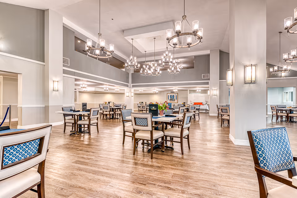 Spacious dining room with multiple wooden tables and chairs featuring blue patterned upholstery. The room has high ceilings with modern chandeliers and wall sconces providing warm lighting. The floor is wooden, and the walls are painted in neutral tones. There is a large mirror on the right side reflecting part of the room.