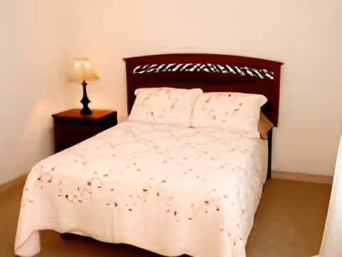 A bedroom with a double bed covered in a light-colored floral bedspread. There is a wooden headboard and a nightstand with a lamp on the left side of the bed. The walls are plain and light-colored.