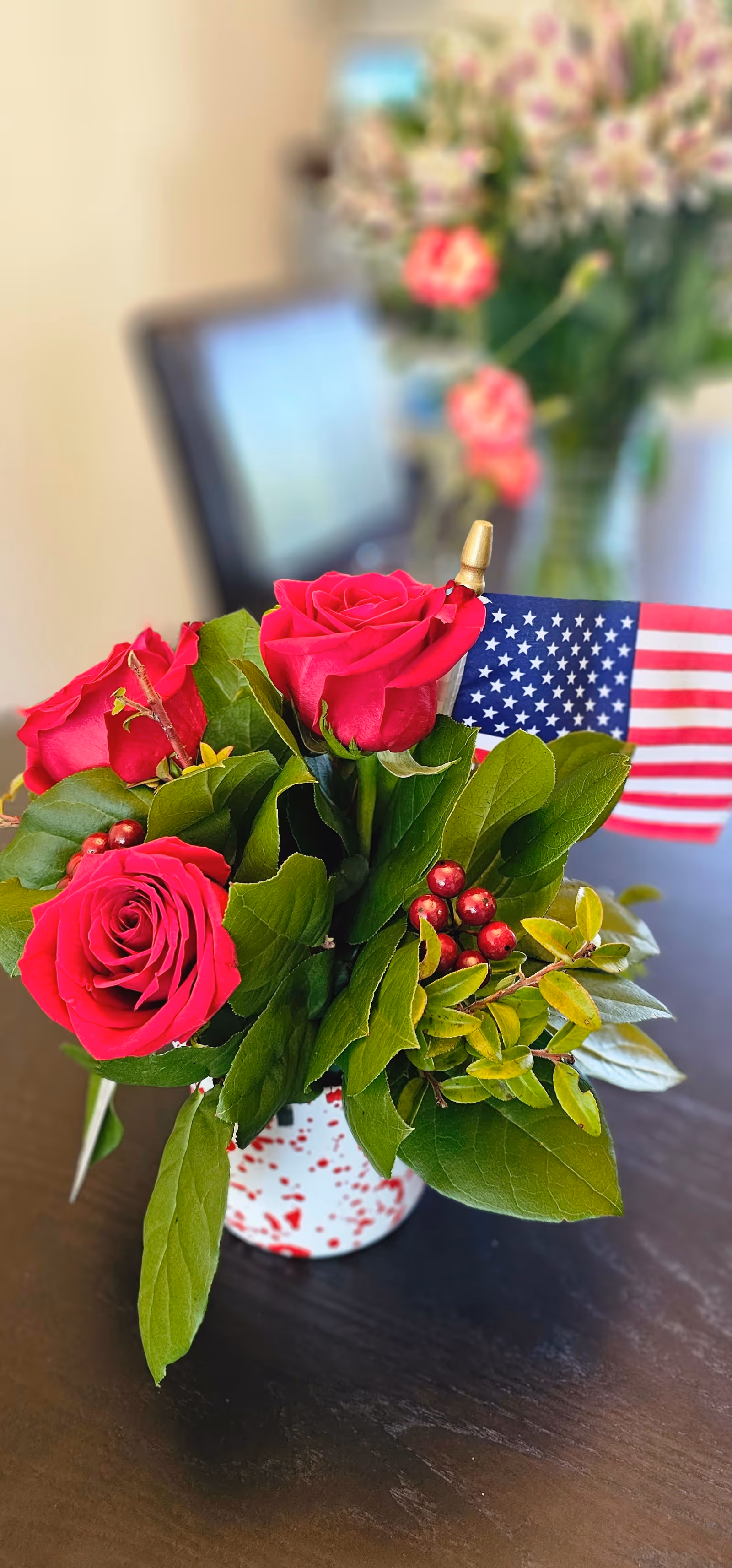 A small vase of red roses and greenery with a miniature American flag on a dark table, with a blurred interior background.