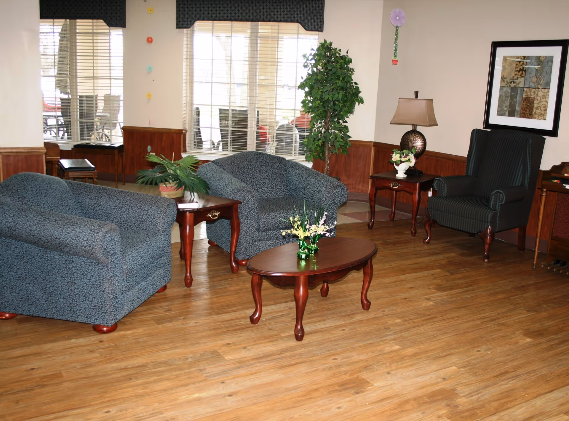A cozy lounge with upholstered armchairs, wooden coffee and side tables, a lamp and potted plants in front of large windows.