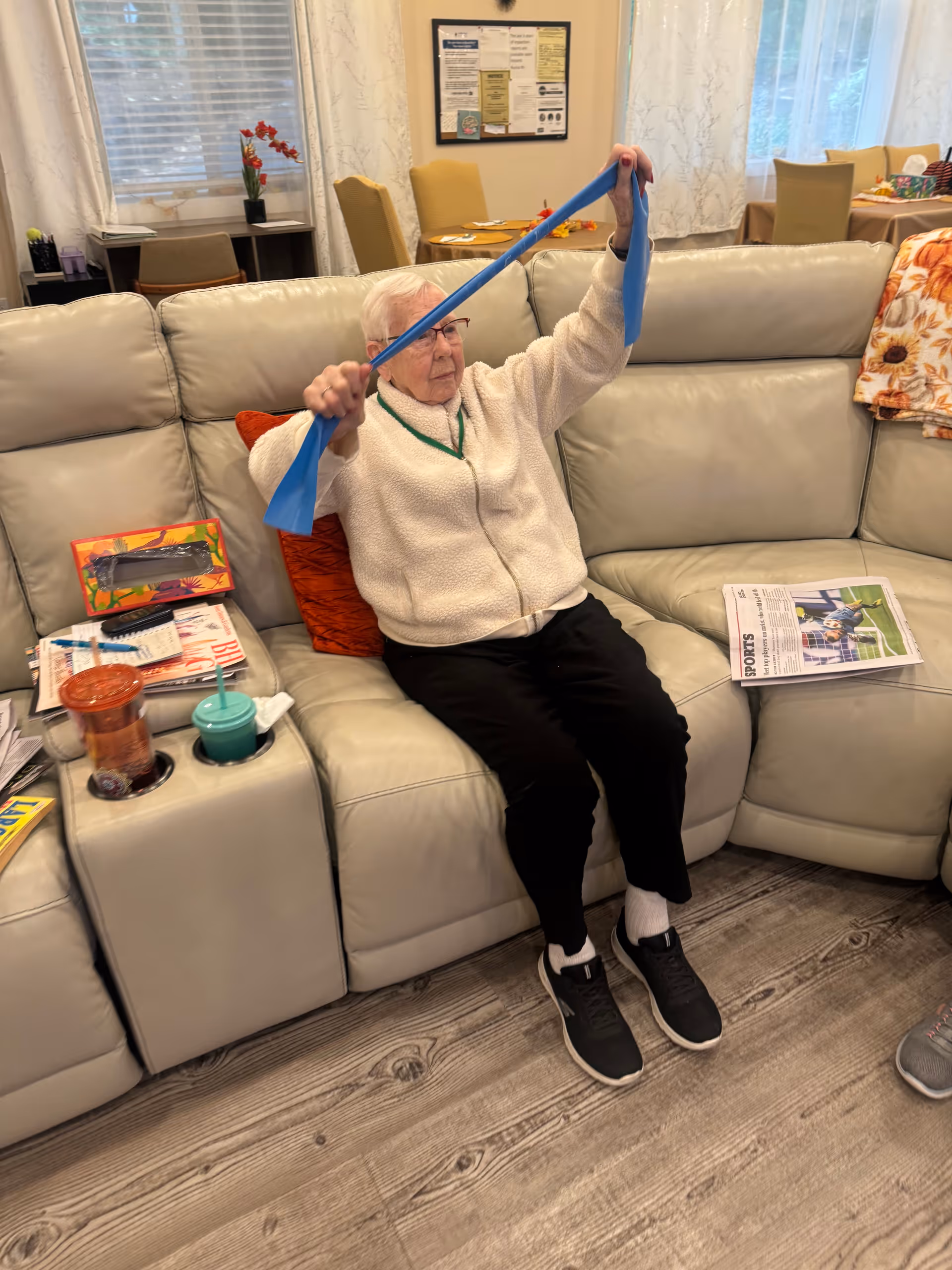 An elderly woman sitting on a light-colored leather couch in a living room, stretching a blue resistance band above her head. There are magazines, a newspaper, and drinks on the armrest next to her. The room has wooden flooring, large windows with curtains, and a dining area with chairs and tables in the background.
