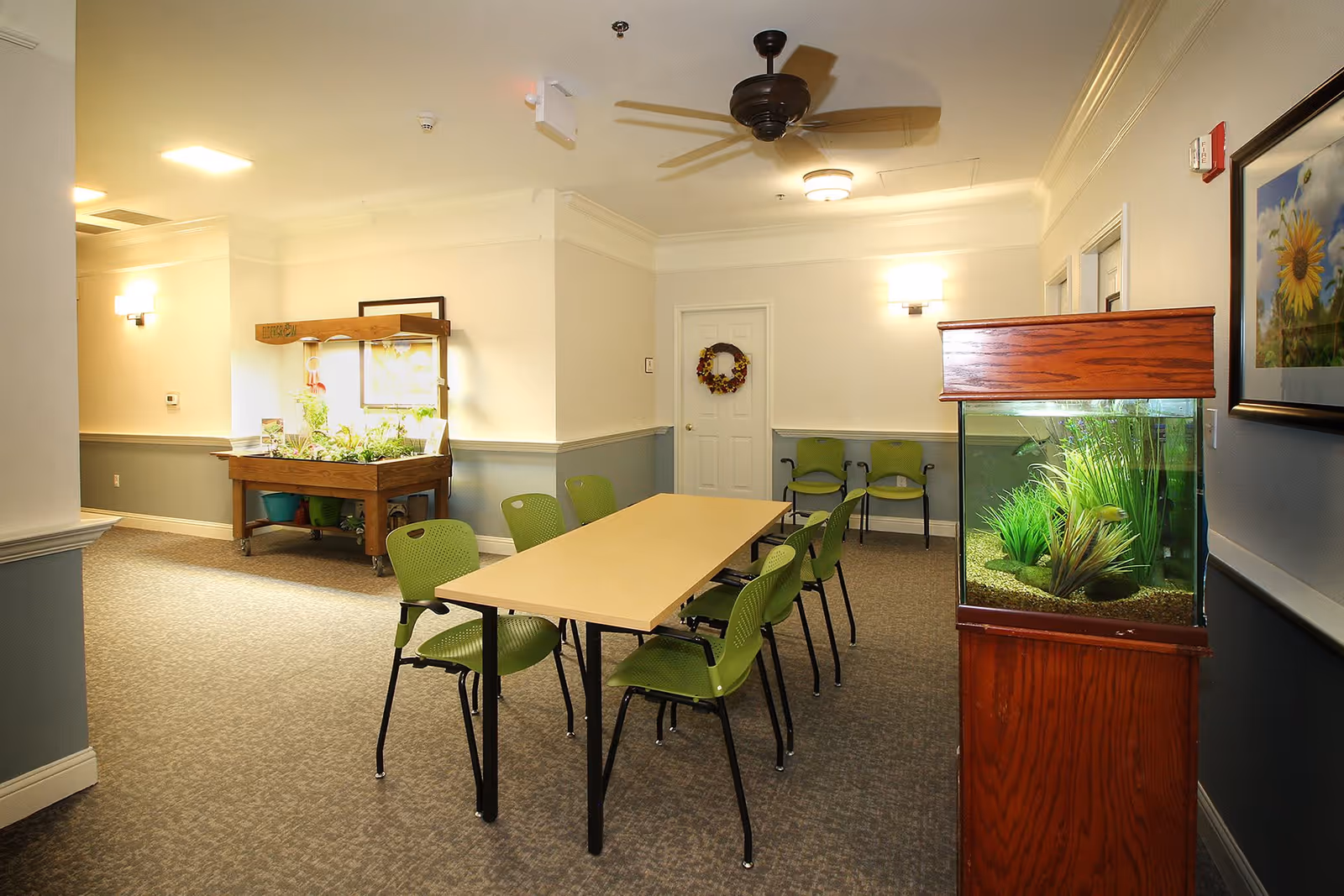 A well-lit common area with a rectangular table surrounded by green chairs. There is a fish tank with plants on the right side and a small indoor garden with plants under a light on the left side. The walls are painted white and light blue with framed artwork and a door decorated with a wreath in the background.