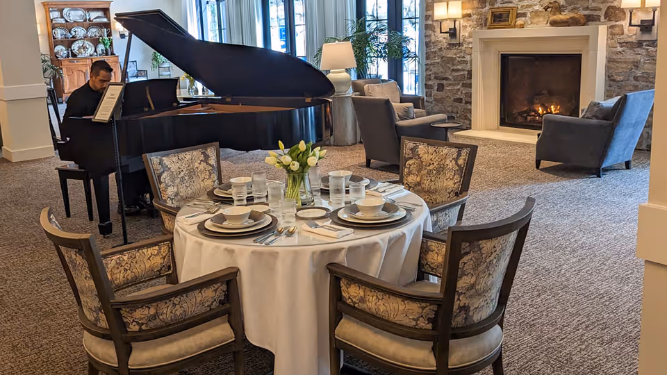 A cozy living room area with a round dining table set for four with plates, bowls, glasses, and silverware. A vase with white tulips is in the center of the table. In the background, a man is playing a grand piano near large windows. There are upholstered chairs and a stone fireplace with a fire burning, creating a warm and inviting atmosphere.