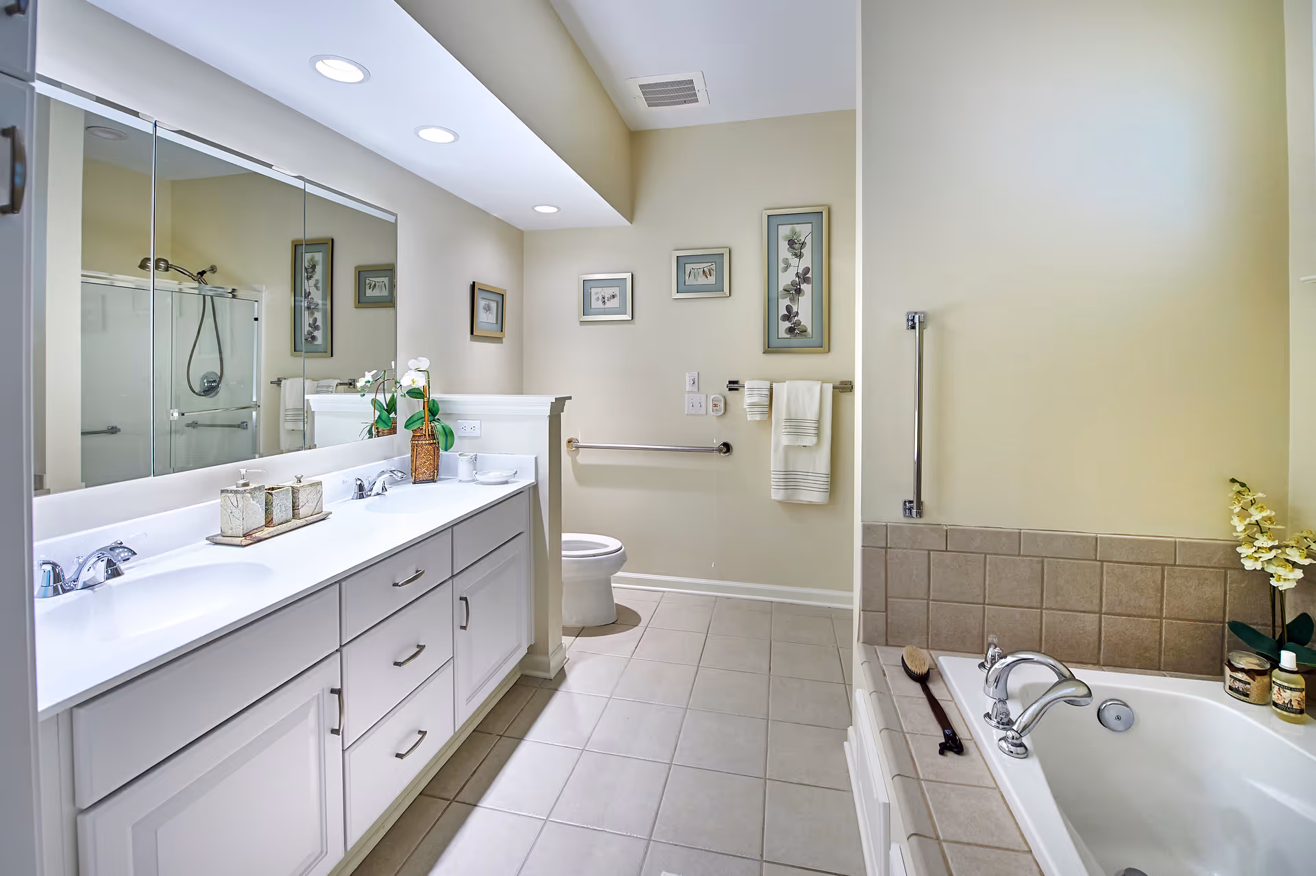 Bright and clean bathroom with a double sink vanity, large mirror, toilet, and a bathtub with tiled surround. The walls are decorated with framed botanical prints, and there are towels hanging on a rail. The floor is tiled, and there is a small plant on the vanity and a flower arrangement near the bathtub.