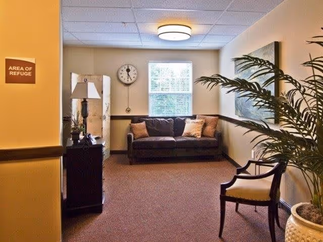 A cozy sitting area in a senior living facility with a dark sofa adorned with pillows, a wooden chair with a cushioned seat, a tall potted plant, a side table with a lamp and phone, a wall clock, and a window letting in natural light. A sign on the wall reads 'AREA OF REFUGE'.