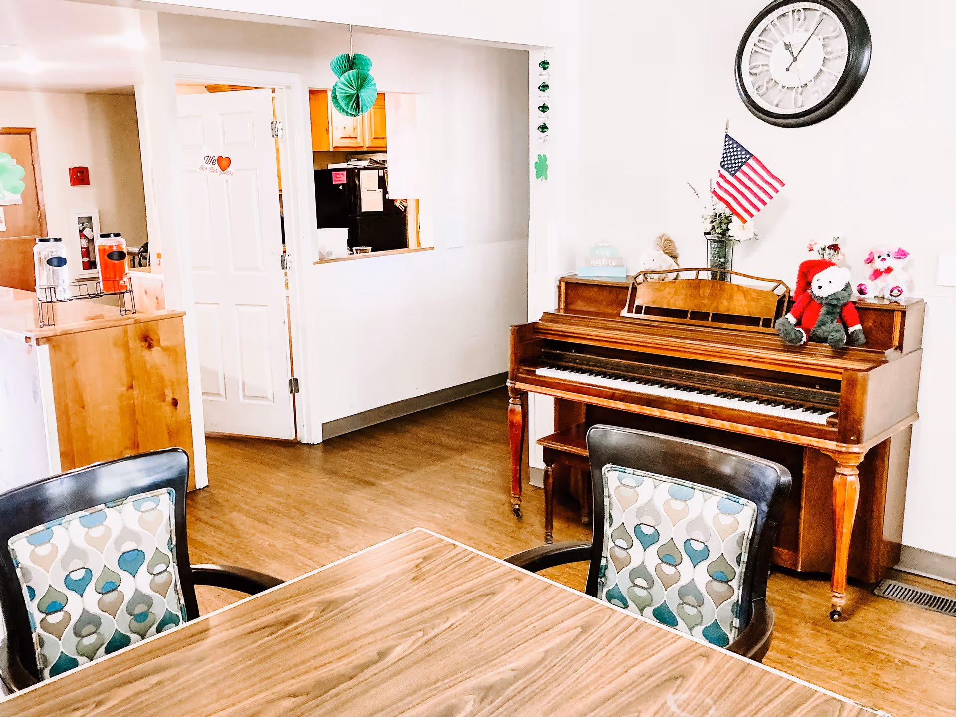 Interior of a senior living facility room with a wooden piano against the wall decorated with stuffed animals and an American flag. A large clock hangs above the piano. In the foreground, there is a wooden table with two chairs that have patterned cushions. The room has wooden flooring and a doorway leading to a kitchen area with wooden cabinets and a black refrigerator. Green paper decorations hang from the ceiling and wall.