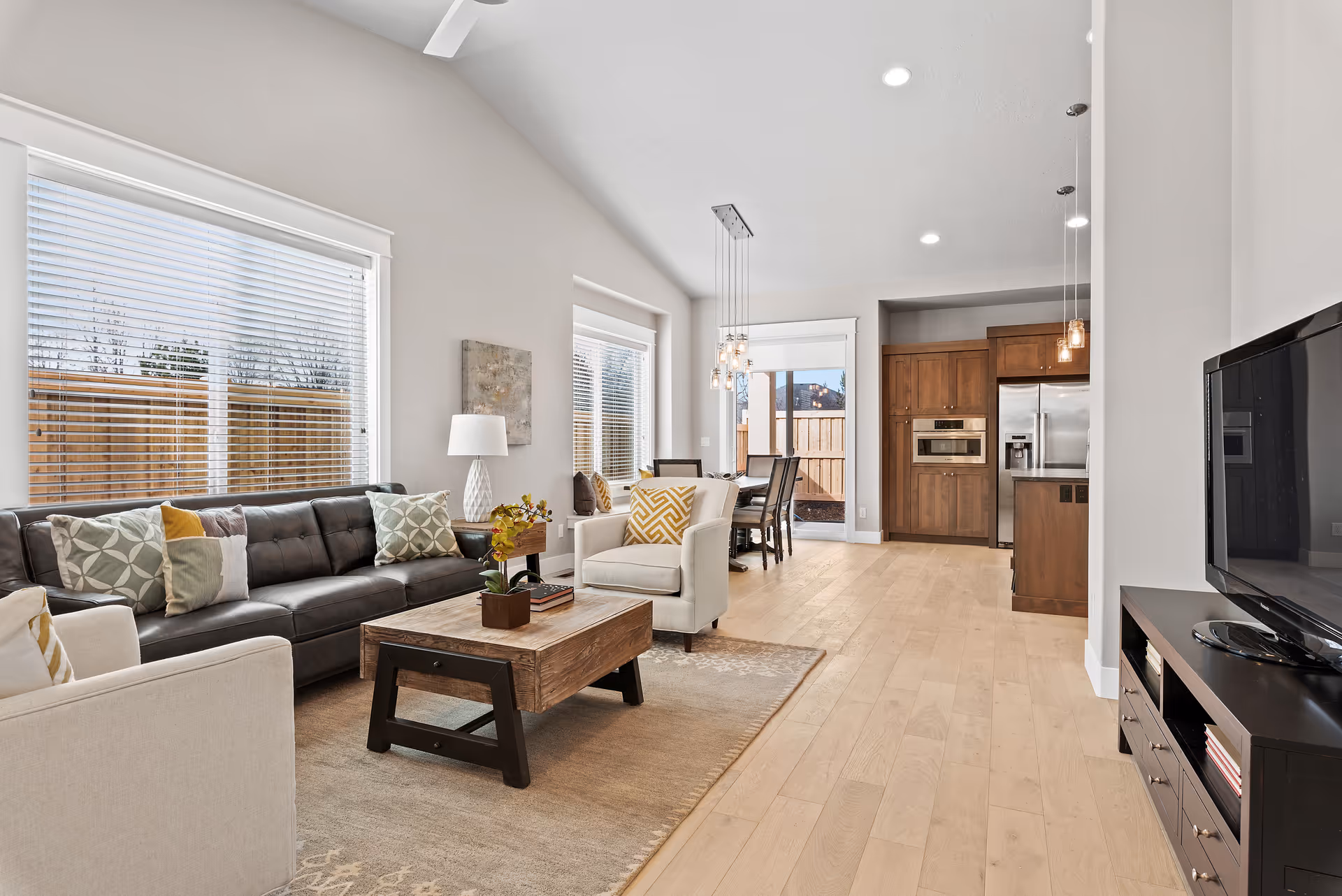 A bright and modern open-concept living room and kitchen area in a senior living facility. The living room features a dark leather sofa with patterned cushions, two beige armchairs, a wooden coffee table with a small plant and books, and a large window with blinds. The kitchen area has wooden cabinets, a stainless steel refrigerator, built-in microwave, and a dining table with chairs under hanging pendant lights. Light wood flooring and neutral-colored walls create a warm and inviting atmosphere.
