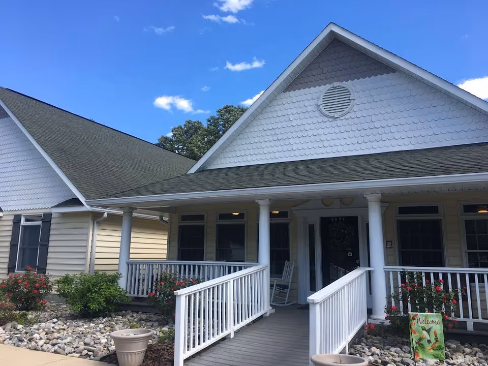 Front exterior view of Peartree House Assisted Living showing a single-story building with a covered porch, white railings, a ramp, rocking chairs, and landscaping with bushes and flowers under a blue sky with a few clouds.