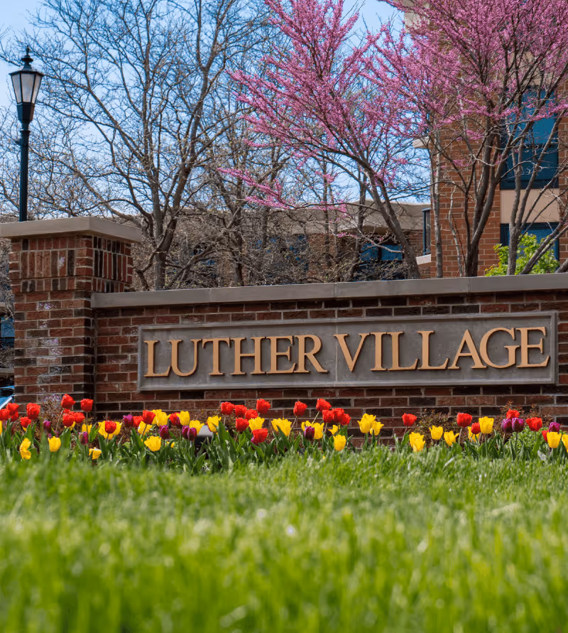 Brick sign with the text 'LUTHER VILLAGE' surrounded by colorful tulips and green grass, with trees and part of a building in the background.