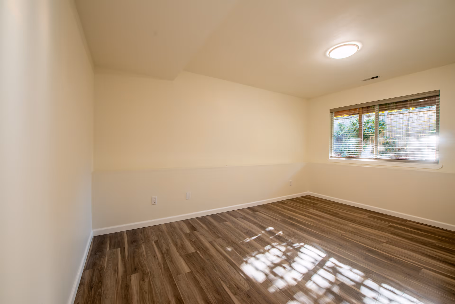 Empty room with light beige walls, wood-patterned flooring, a window with blinds partially open showing greenery outside, and a ceiling light fixture.