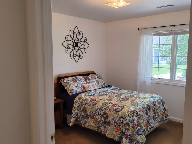 A small bedroom with a bed covered in a colorful patterned quilt and pillows. There is a wooden headboard and a nightstand beside the bed. A decorative metal wall art piece hangs above the bed. A window with sheer curtains lets in natural light and shows a green lawn outside.