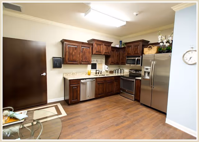 A modern kitchen with dark wooden cabinets, stainless steel appliances including a refrigerator, oven, microwave, and dishwasher. The kitchen has a light-colored granite countertop and wood flooring. There is a dark brown door on the left side and a glass table partially visible in the foreground.