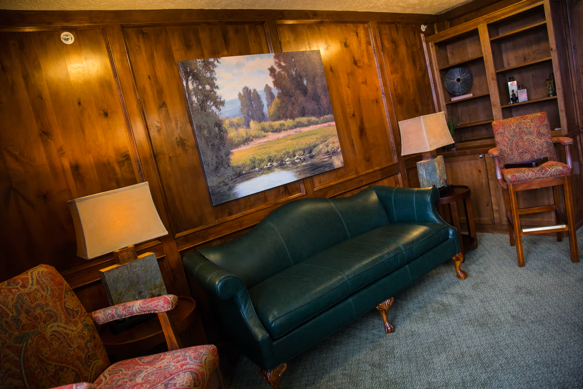 A cozy room with wood-paneled walls featuring a green leather sofa, two patterned armchairs, two table lamps on small round tables, a built-in wooden bookshelf, and a landscape painting hanging above the sofa.