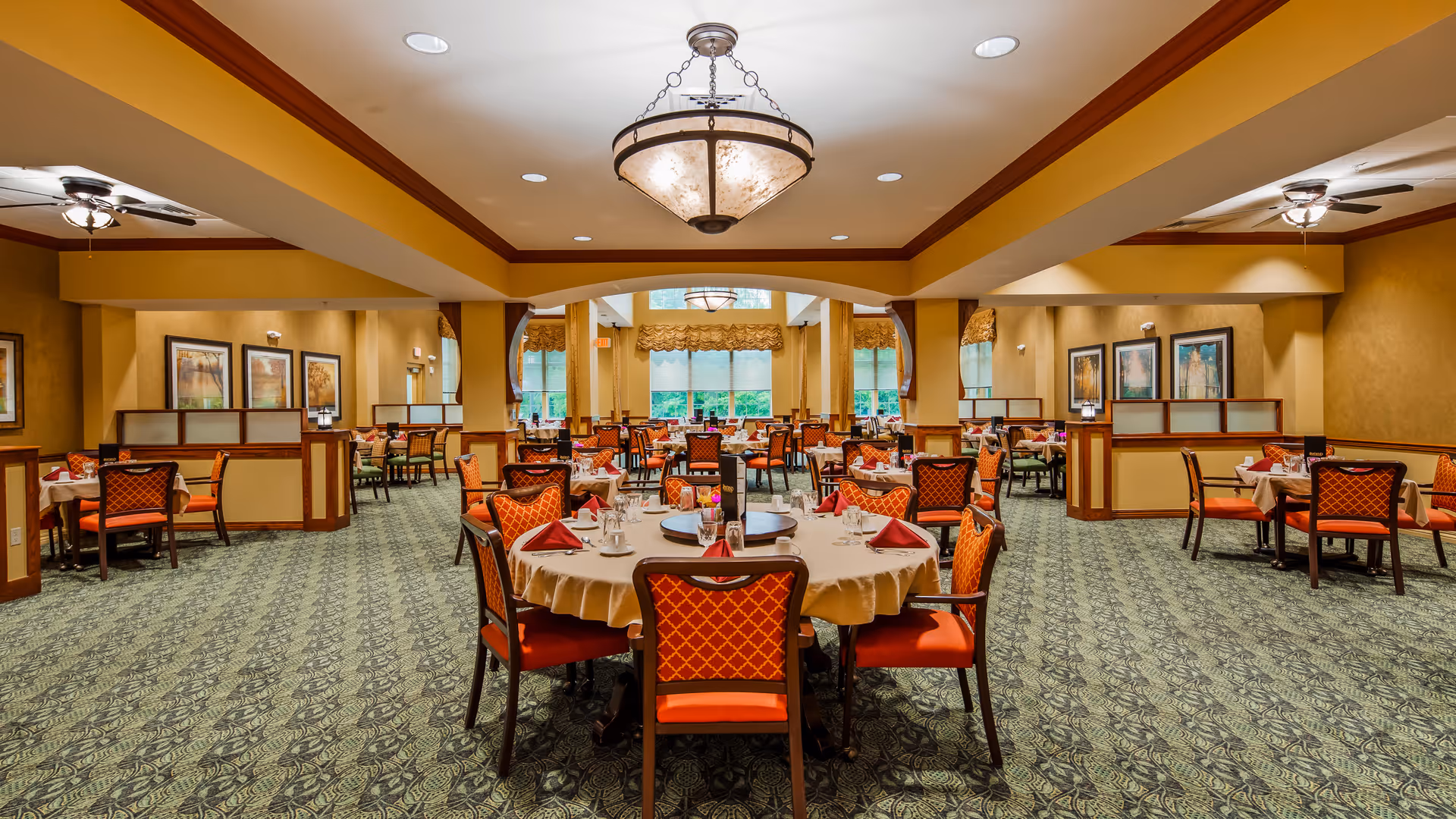 A spacious dining room with round tables covered with beige tablecloths and set with red napkins, glassware, and silverware. The room features patterned carpet, warm yellow walls, framed artwork, and large windows letting in natural light. Elegant ceiling lights and ceiling fans illuminate the space.