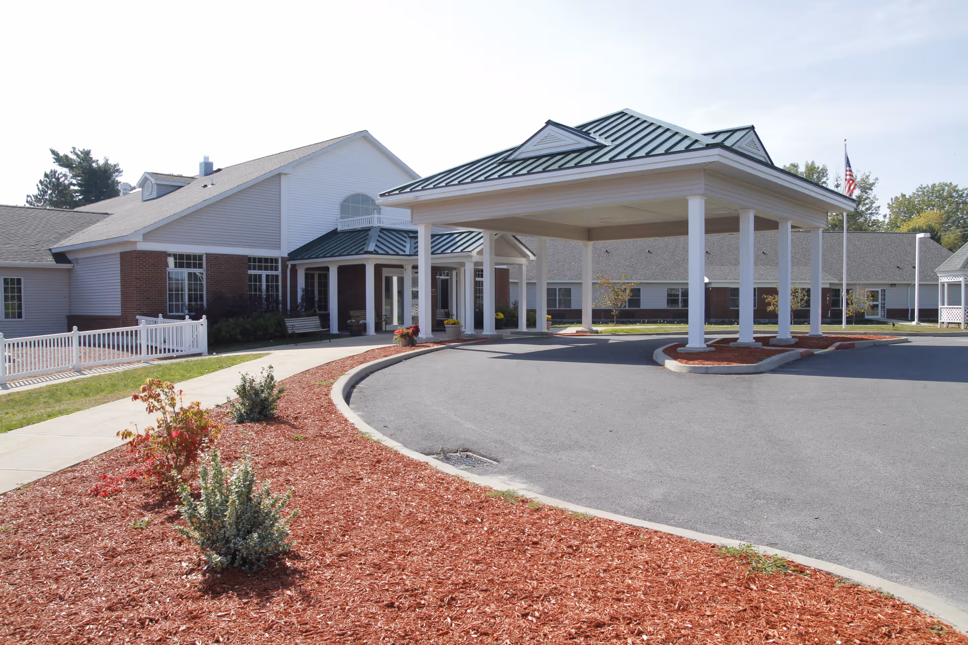 Exterior view of Pine Harbour senior living facility showing a covered entrance with white pillars, a circular driveway, landscaped areas with mulch and small shrubs, and a building with white siding and brick accents under a clear sky.