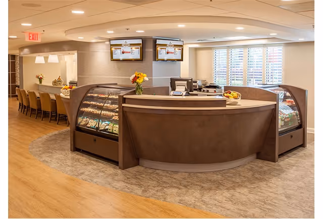 Interior view of a modern reception or service counter area in a senior living facility with curved brown counters displaying snacks and fruits. Behind the counter are two monitors mounted on the wall, a cash register, and a vase with flowers. To the left, there is a seating area with chairs and a counter under pendant lights. The floor has a combination of wood and carpet, and windows with white blinds allow natural light in.