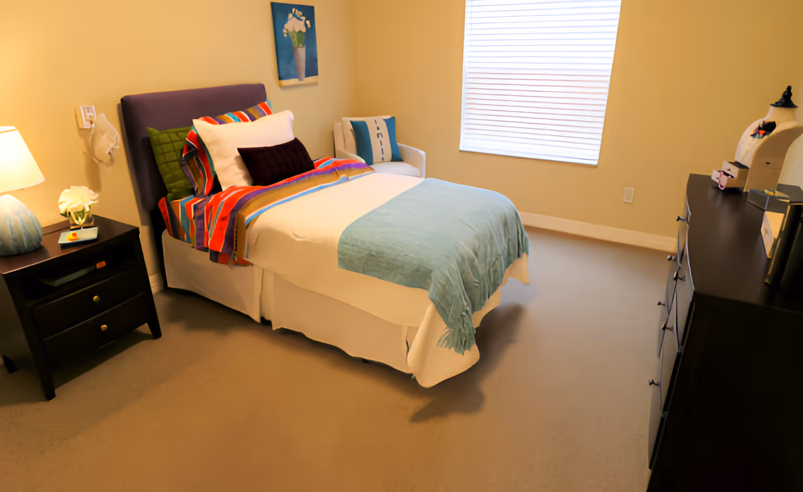 Small furnished bedroom with a neatly made twin bed covered by colorful pillows and throws, a nightstand with a lamp, dresser, and a window with blinds.