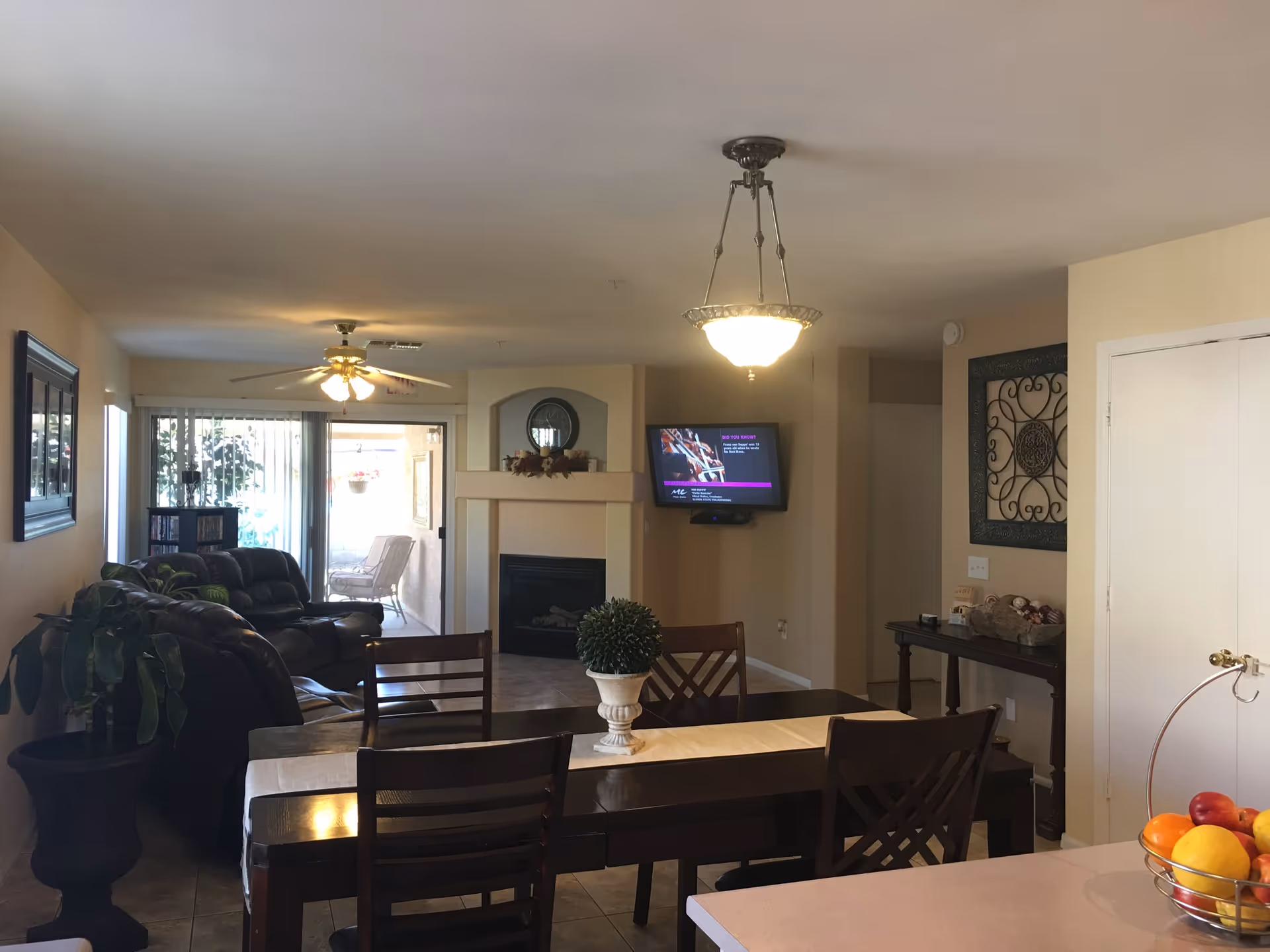 Interior view of a senior living facility showing a dining area with a dark wooden table and chairs, a small decorative plant centerpiece, a living room with leather sofas, a ceiling fan, a wall-mounted TV above a fireplace, and a sliding glass door leading to an outdoor patio with chairs. There is also a side table with decorative items and a fruit basket on a countertop in the foreground.