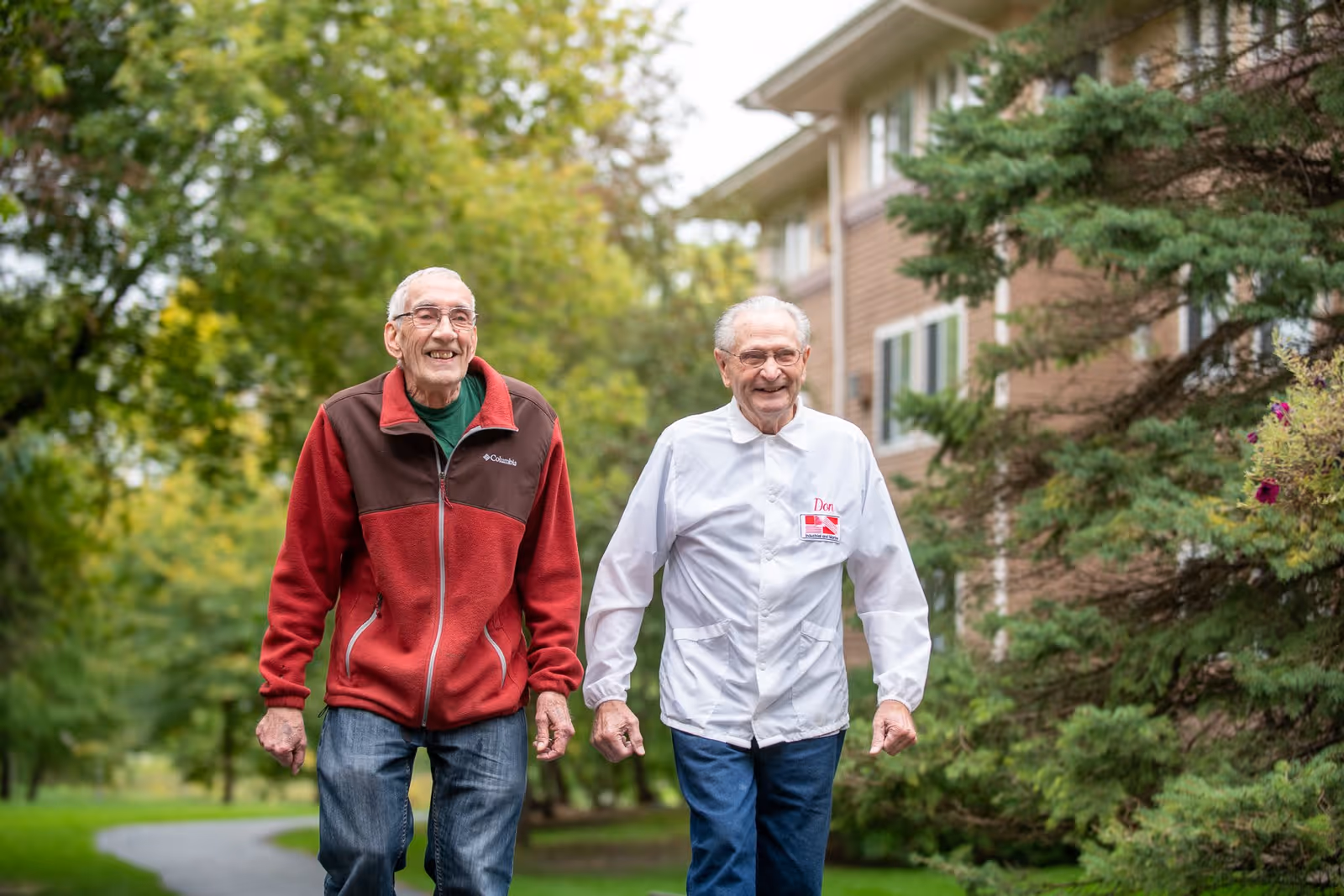 Two elderly men walking outdoors on a paved path surrounded by green trees and grass, with a multi-story building visible in the background. Both men are smiling and appear to be enjoying the walk.