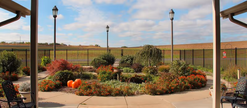 A landscaped outdoor garden area with various shrubs, flowers, and pumpkins arranged in a circular pattern. The garden is surrounded by a paved walkway and black metal fencing. There are three tall street lamps and a couple of black metal chairs under a covered patio. The sky is partly cloudy with blue patches.