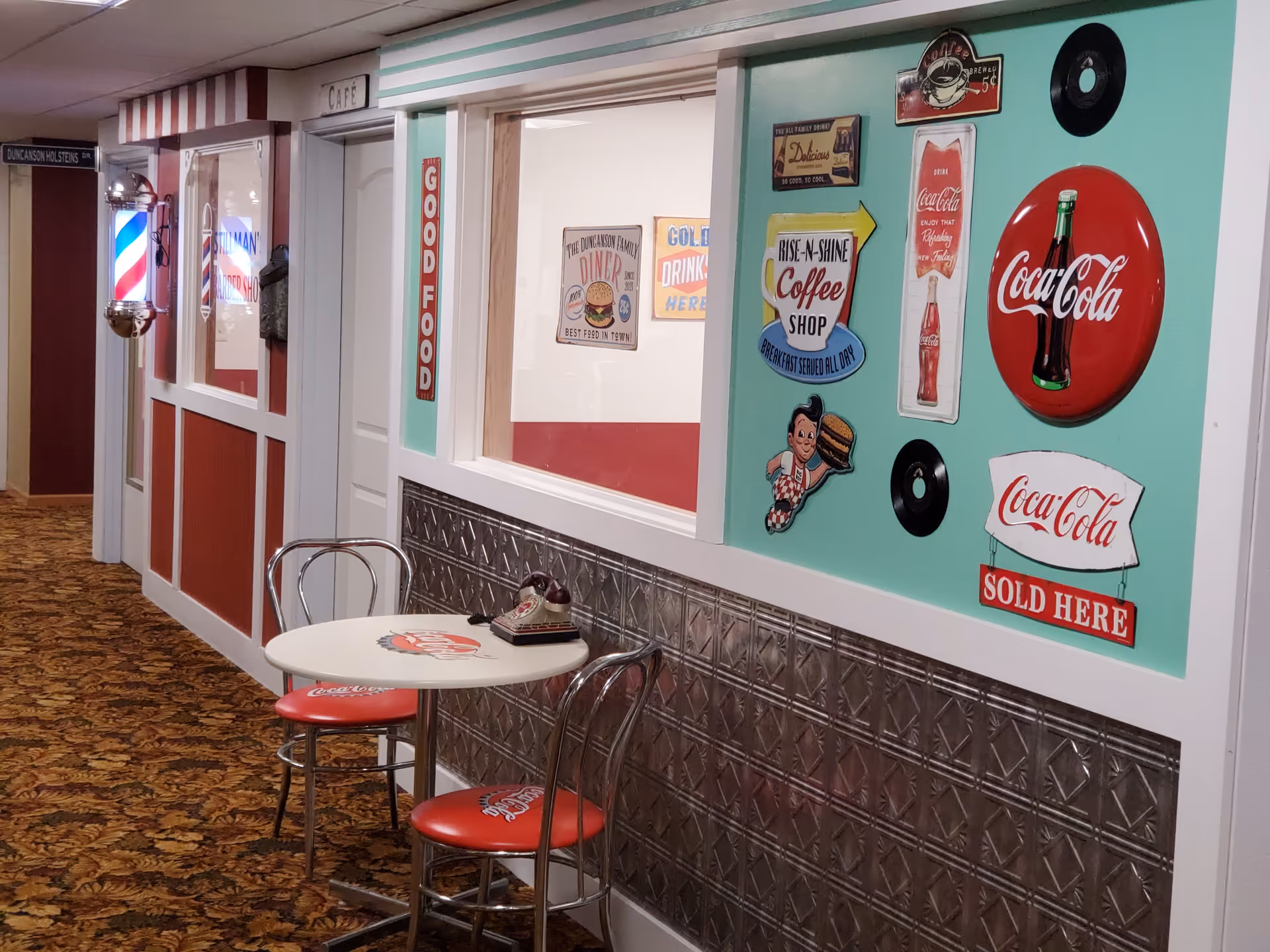 A hallway in Lewiston Senior Living decorated with vintage diner and Coca-Cola themed signs. There is a small round table with two red Coca-Cola cushioned chairs and an old-fashioned rotary phone on the table. The walls have retro diner-style decor including a barber shop sign and various nostalgic advertisements.