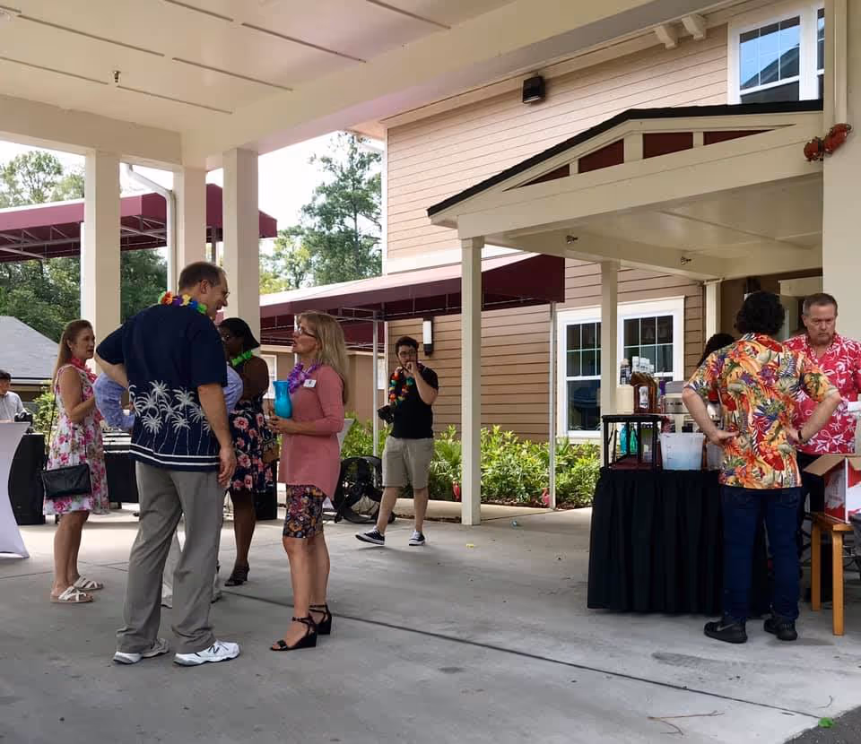 A group of people socializing outdoors under a covered patio area at a senior living facility. Some individuals are wearing colorful Hawaiian shirts and leis, and there is a table with drinks and supplies set up nearby. The background shows the exterior of the facility building with windows and greenery.