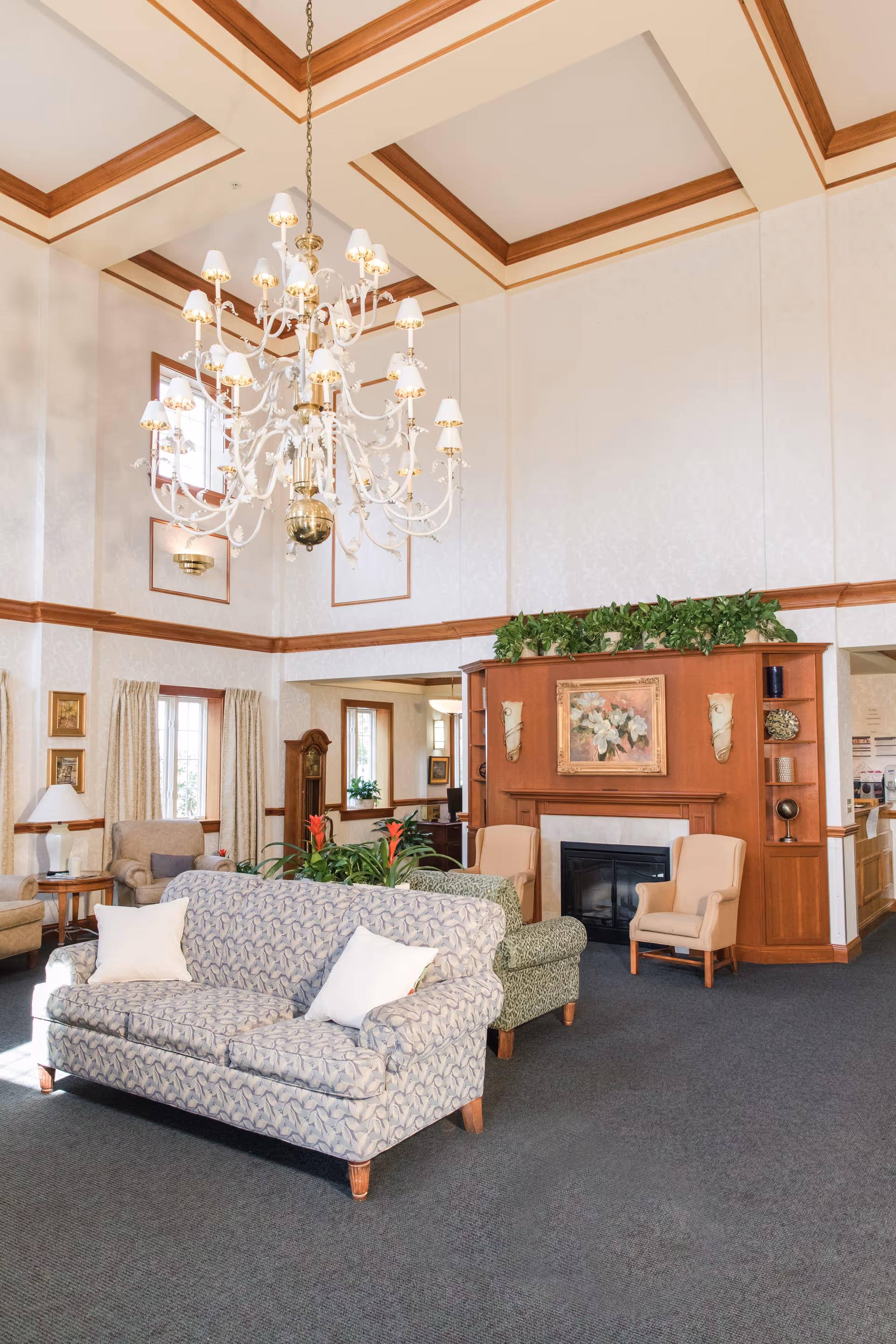 A bright and spacious living room area in a senior living facility featuring a patterned sofa with white pillows, two beige armchairs, a wooden fireplace mantel with a floral painting above it, and a large ornate chandelier hanging from a high ceiling with wooden trim. There are windows with curtains, plants on the mantel and in the room, and a carpeted floor.